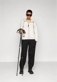 Bel plašč iz flisa v beli barvi z zadrgo spredaj in kapuco, kombiniran s črnimi cargo hlačami in črnimi pohodniškimi čevlji, drži pohodniški palico.