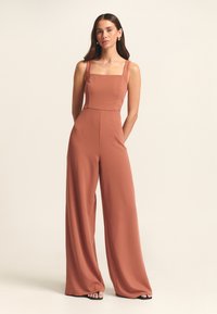 Roestkleurige jumpsuit met vierkante halslijn, brede pijpen en dikke bandjes. Gemaakt van een zachte stof, heeft het een aansluitende taille en minimale naden.