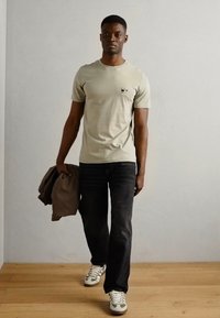 Gai bēšas īsām piedurknēm T-krekls ar logotipa detaļām, apvienots ar tumšajām džinsām un sporta apaviem ar zaļiem akcentiem. Modelis tur brūnu džemperi.