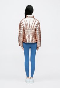 Fémes rózsaarany steppelt dzseki, testhezálló derékkal, hátulnézetben. Kék leggingsszel és fehér tornacipővel párosítva. Simább textúra.