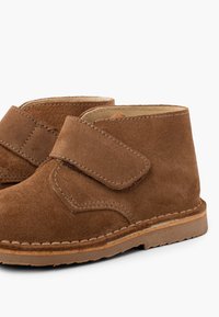 Bottes chelsea en daim marron avec une sangle en velcro, des accents piqués et une semelle en caoutchouc beige durable. Forme arrondie et design minimaliste.