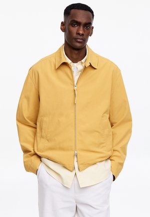 Uomo che indossa una giacca giallo senape con zip sopra una camicia crema con bottoni e pantaloni bianchi, in piedi con le mani nelle tasche della giacca.