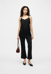 Γυναίκα με κατσαρά μαλλιά που φοράει μαύρο καμιζόλ με δαντέλα, μαύρο παντελόνι, μαύρες γόβες με χαμηλό τακούνι, κρατώντας καφέ δερμάτινη τσάντα.