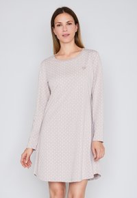 Donna che indossa un vestito lungo rosa chiaro, a maniche lunghe, al ginocchio, con un sottile motivo geometrico, in piedi contro uno sfondo chiaro e semplice.