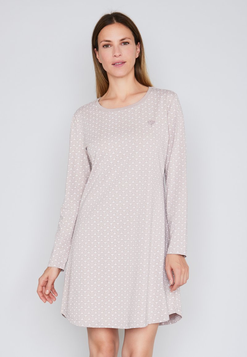 Donna che indossa un vestito lungo rosa chiaro, a maniche lunghe, al ginocchio, con un sottile motivo geometrico, in piedi contro uno sfondo chiaro e semplice.