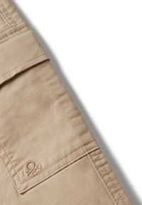 Bežová tkanina zblízka s viditelným švem a malou vyšitou nášivkou blízko manžety rukávu oděvu nebo nohavice.