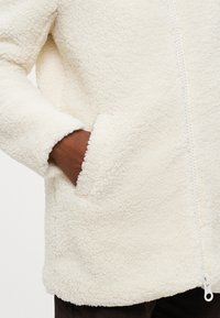 Veste en polaire couleur crème avec fermeture éclair à l'avant, tissu texturé et poches latérales. Matériau doux et moelleux avec des poignets roulés.
