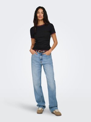 Kvinne med langt mørkt hår som har på seg en svart t-skjorte med mørkerød kant, lyseblå vide jeans, brunt belte og brune joggesko, stående med hendene i lommene.