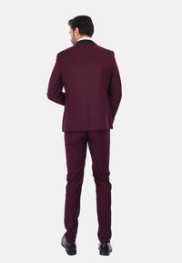 Abito bordeaux con vestibilità su misura. Presenta un rever a punta nero, pantaloni coordinati e scarpe eleganti nere lucide. Tessuto con una consistenza morbida.