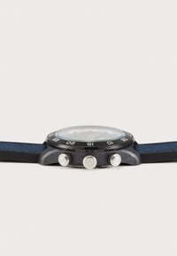 Montre chronographe avec un grand boîtier rond noir, un verre transparent et un bracelet en tissu bleu foncé. Trois boutons sur le côté pour la fonctionnalité.