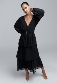 Schwarzes Chiffonkleid mit Lagen-Design, tiefem V-Ausschnitt und langen, weiten Ärmeln. Ein goldenes Gürtel-Detail betont die Taille. Kombiniert mit goldenen Riemchenhighheels.