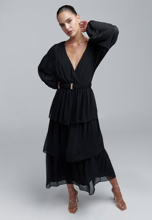 Schwarzes Chiffon-Maxikleid mit Glockenärmeln, gestuftem Design, tiefem V-Ausschnitt, elastischem Taillengummi und goldener Schnallenakzent. Getragen mit goldenen Riemchenabsätzen.