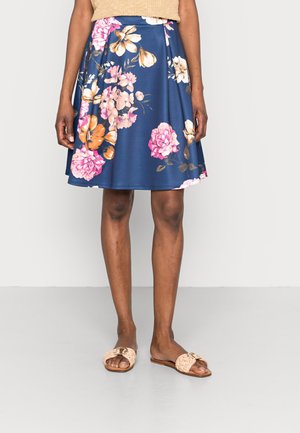 Mujer con falda azul marino hasta la rodilla con grandes estampados florales en rosa, naranja y blanco, top beige de punto y sandalias beige tejidas deslizantes.