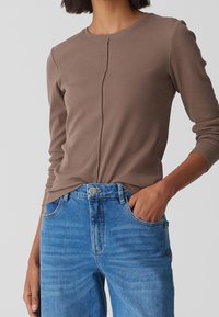 Kvinna iklädd en tät långärmad brun topp som är stoppad i högmidjade blå jeans, med en hand i fickan, stående mot en enkel bakgrund.