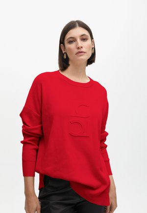 Red knitted sweatshirt with a crew neckline and oversized fit. Features a raised logo design on the front and ribbed texture at cuffs.