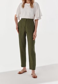 Høj talje olivengrønne linnedbukser med lige ben, sidelommer og subtil struktur, parret med en hvid bluse med volanger og sandaler.