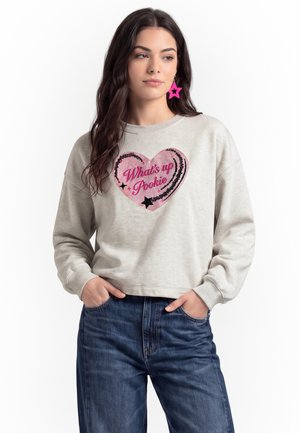 Donna con lunghi capelli scuri che indossa una felpa grigio chiaro con un cuore rosa e il testo "What's up Pookie" e orecchini a stella rosa, mani nelle tasche.