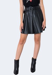 Jupe plissée en similicuir noir avec taille ceinturée. Portée avec des bottines noires ajustées à la cheville. Texture lisse et silhouette évasée.