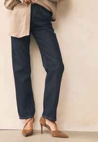 Tamsiai mėlynos spalvos džinsai iš džinso audinio, tiesiomis kojinėmis, kontrastingu šviesiu siuvimu. Dėvimi su stilizuotais rudos spalvos zomšos aukštakulniais batais.