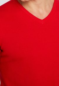 Maglione rosso in cotone con scollo a V, caratterizzato da un design aderente e orlo a coste, che mostra una texture liscia e un colore vivace.