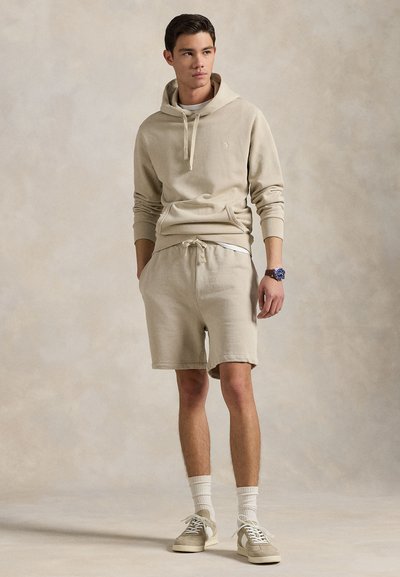 Lys beige hettegenser med snorer og kengurulomme, kombinert med matchende shorts. Modellen har på seg hvite sokker og lyse joggesko.