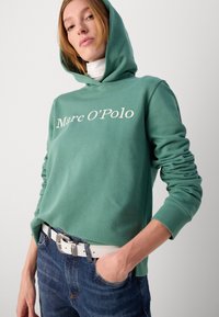 Felpa verde realizzata in morbido tessuto, caratterizzata da un ampio logo bianco "Marc O'Polo" davanti. Abbinata a jeans blu scuro e una cintura bianca.