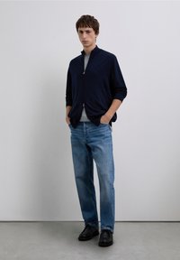 Granatowy sweter na zamek błyskawiczny nałożony na szarą koszulkę, w połączeniu z jasnoniebieskimi dżinsami i czarnymi butami, stojący na neutralnym tle.