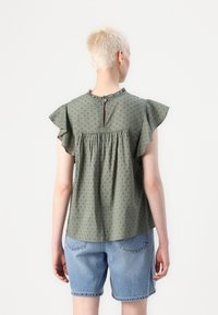 Grøn bluse med tekstur, rynkede ærmer og knaplukning bagpå, parret med lyse denimshorts.
