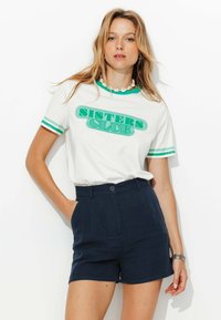 T-shirt en coton blanc avec un graphique "SISTERS CLUB" vert, col vert et rayures aux manches vertes, associé à un short taille haute bleu marine foncé.