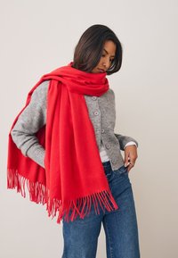 Écharpe en laine rouge aux bords frangés, drapée sur un cardigan gris. Texture douce et couleur vive contrastant avec la tenue neutre.