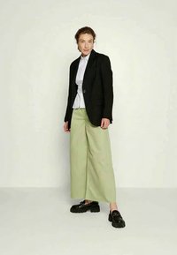 Blazer preto, camisa branca com nó, calças largas verde-claras e sapatos pretos chunky. Texturas suaves com corte ajustado.