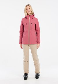 Rosa wasserdichte Jacke mit Kapuze, ausgestattet mit zwei Reißverschlusstaschen auf der Brust und Belüftungslöchern. Kombiniert mit beigen Skihosen und schwarzen Stiefeln.