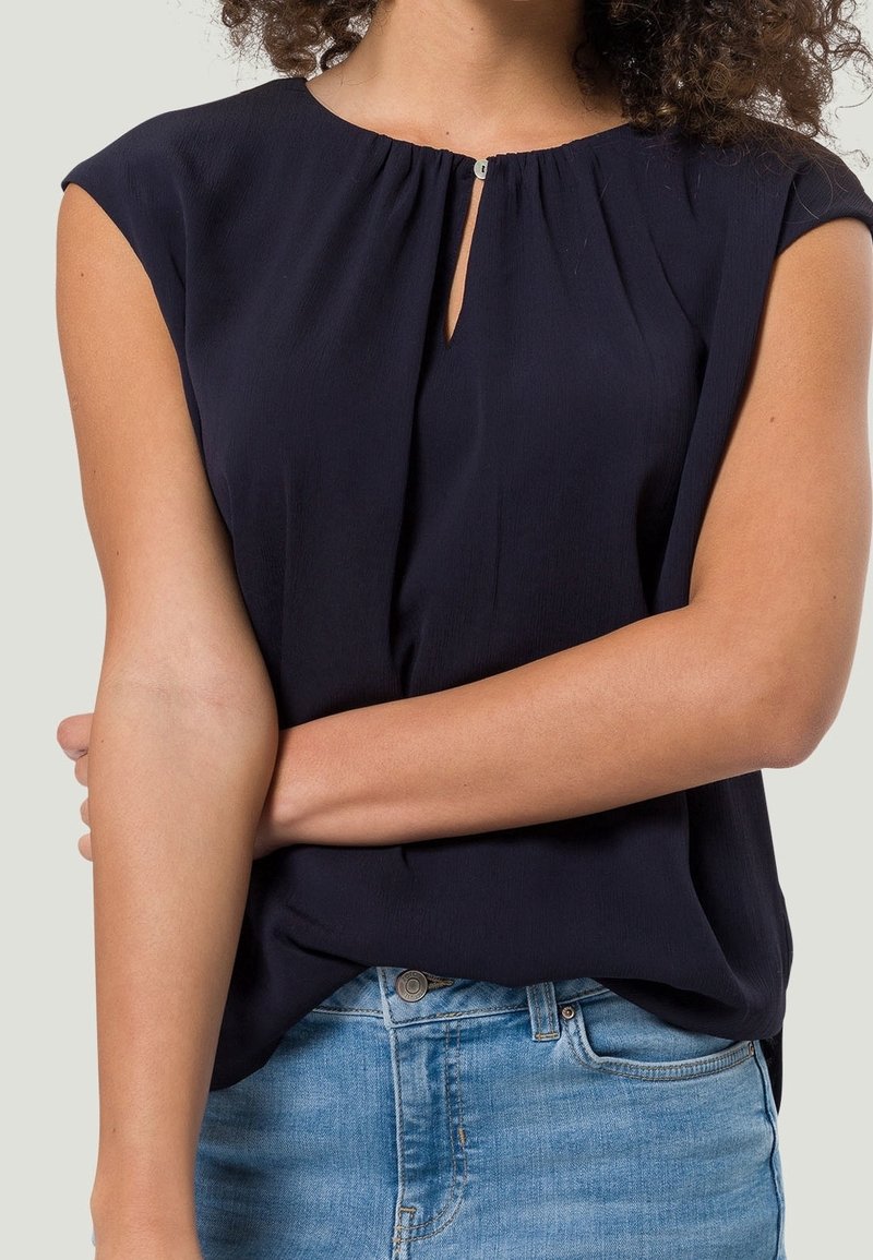 Top blu navy a maniche corte realizzato in tessuto morbido, con scollatura a chiave e dettagli arricciati, abbinato a jeans in denim azzurro chiaro.