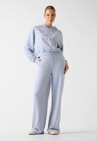 Jasnoniebieska bluza z kapturem z wyszywanym logo, zestawiona z dopasowanymi, szerokimi spodnimi o wysokim stanie. Gładka tkanina, luźny krój.