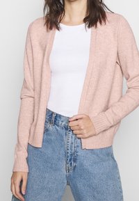 Ljusrosa stickad cardigan med långa ärmar, öppen fram och ribbade muddar, som är parat med hög midja blå denimjeans.