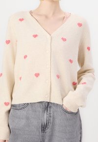 Krämfärgad cardigan med V-urringning, försedd med spridda rosa hjärtmönster, sex knappar och ribbade ärmslut, ihopparad med grå högmidjade byxor.