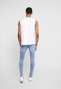 Vit ärmlös skjorta i kombination med ljusblå skinny jeans och vita sneakers. Bakgrunden är enkel och neutral.