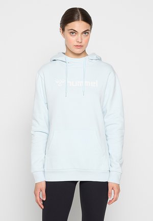 Kobieta z brązowymi włosami, ubrana w jasnoniebieską bluzę z kapturem marki hummel i czarne spodnie, stoi na tle jasnego, jednolitego tła.