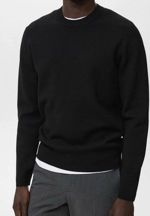 Man draagt een zwart lange-mouwen trui met ronde hals over een wit shirt, gecombineerd met grijze broek tegen een effen achtergrond.