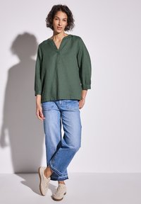 Tunika di lino verde con scollo a V e maniche a tre quarti, abbinata a jeans a gamba larga blu chiaro e sneakers beige testurizzate.