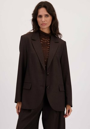 Vrouw met lang, donker haar, gekleed in een diepbruine kanttop en bijpassende oversized blazer en broek, staand tegen een effen achtergrond.