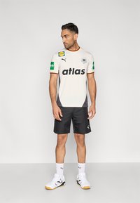 Balts sporta krekls ar melnu "atlas" logotipu, oranžiem akcentiem un zaļām sponsoru uzlīmēm, kopā ar melnām šortiem un baltām sporta kurpēm.