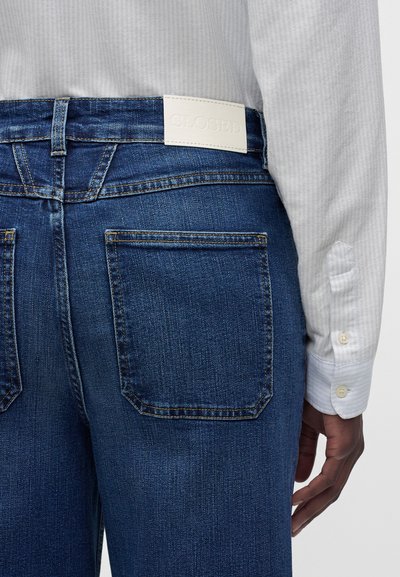 Gros plan d'une personne portant un jean en denim bleu avec des poches arrière et une étiquette blanche, associé à une chemise blanche à manches longues.