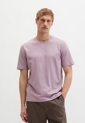 Jonge man met kort blond haar, gekleed in een lavendelkleurig T-shirt met korte mouwen en een ronde hals, en een bruine broek, staand met één hand in zijn zak.