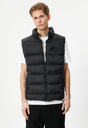 Sort puffervest med høj krave, lynlås foran og et patch-logo på venstre bryst. Har quiltet design og en glat, skinnende tekstur.