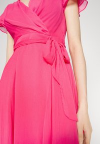 Robe portefeuille rose en tissu texturé, manches courtes à volants et ceinture nouée à la taille. La robe présente un décolleté en V profond et une jupe fluide.