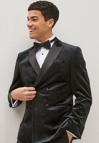Chaqueta de esmoquin de terciopelo negro con solapas de satén, diseño de doble botonadura y dos bolsillos frontales. Llevada con una camisa blanca y una pajarita negra.