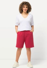 Witte v-hals t-shirt met omgeslagen mouwen, gecombineerd met knielange rode shorts. Model draagt witte sneakers, met handen in de zakken, kroeshaar.