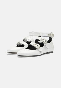 Zapatos de charol blancos con recortes, correas en el tobillo y grandes ojales decorativos. Diseño elegante y estilizado con punta afilada.