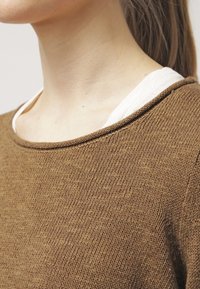 Maglione marrone lavorato a maglia con scollo rotondo, tessuto testurizzato e cuciture a contrasto. Indossato sopra una camicia color crema al colletto.